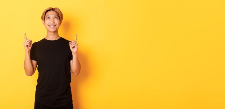 Handsome smiling asian man in black t-shirt pointing fingers up, looking at banner pleased, yellow background.