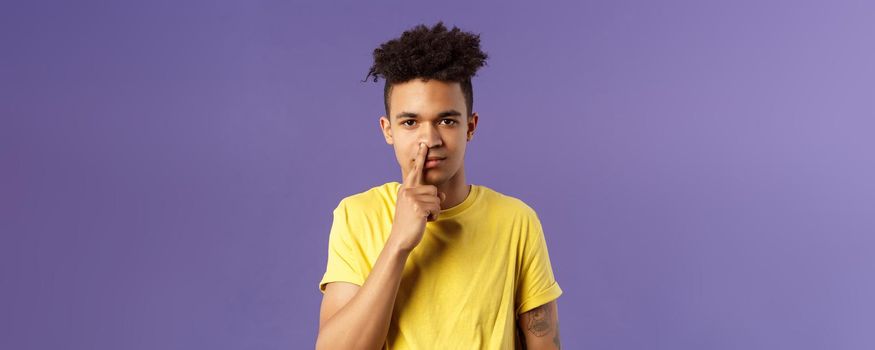 Close-up portrait of funny and unappropriate young hispanic teenage guy picking nose with finger, smiling and looking at camera, standing purple background, have bad habit.