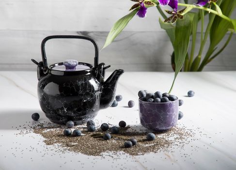 still life with a black ceramic teapot and a cup of tea with blueberries on a light background with an orchid, chia seeds and berries are scattered on the table,High quality photo