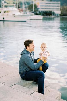 Dad sits on the pier with a baby on his lap against the backdrop of yachts. High quality photo