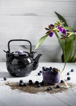 still life with a black ceramic teapot and a cup of tea with blueberries on a light background with an orchid, chia seeds and berries are scattered on the table,High quality photo