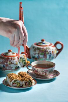 Japanese style tea party with vintage porcelain set, with life motives, sweet rolls dessert for breakfast on a blue background, hand holding chopsticks roll, HIgh quality photo