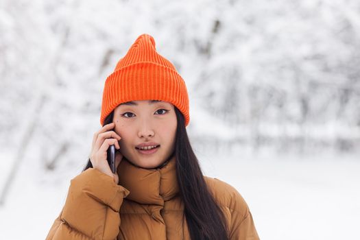 Beautiful asian woman in warm clothes talking on the phone in the park on a winter snowy day on a walk