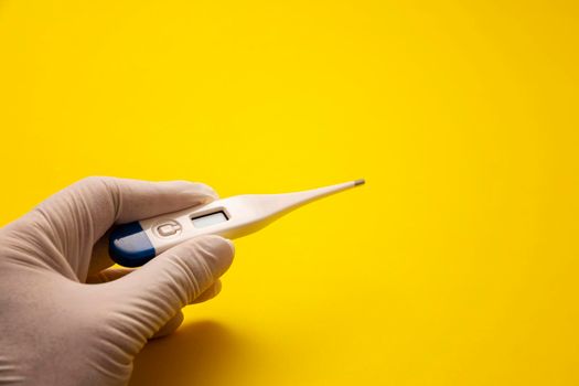 hand with medical latex glove holding a digital thermometer on yellow background. Fever and Healthcare concept. Copy space
