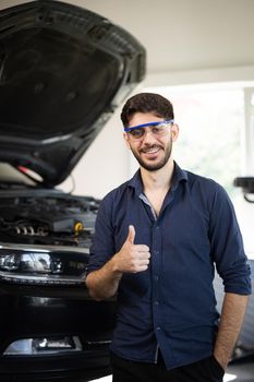 Portrait of young caucasian bearded handsome man auto mechanic or manager in goggles smiling to camera and giving thumb up in garage. Cheerful guy standing at cars service.