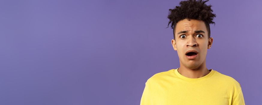 Close-up portrait of shocked, alarmed young man gasping, open mouth scared and frightened, staring camera concerned, facing troublesome shocking news, purple background. Copy space