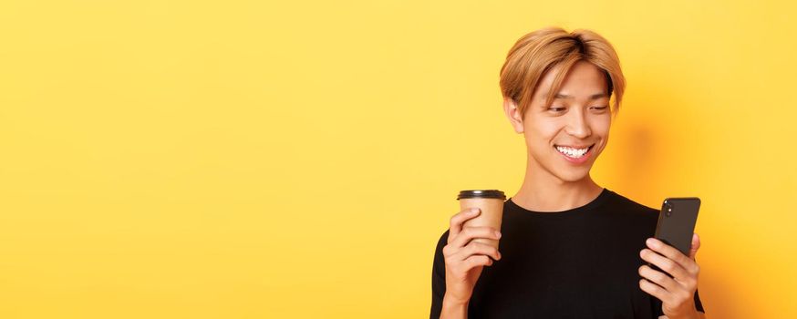 Close-up of handsome stylish asian male student, using mobile phone and drinking coffee. Guy messaging on smartphone, standing over yellow background.