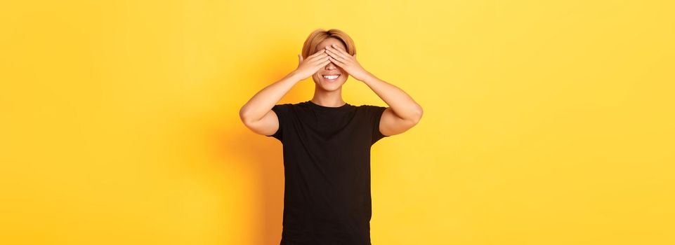 Portrait of excited happy asian guy, smiling and close eyes with hands, standing yellow background.