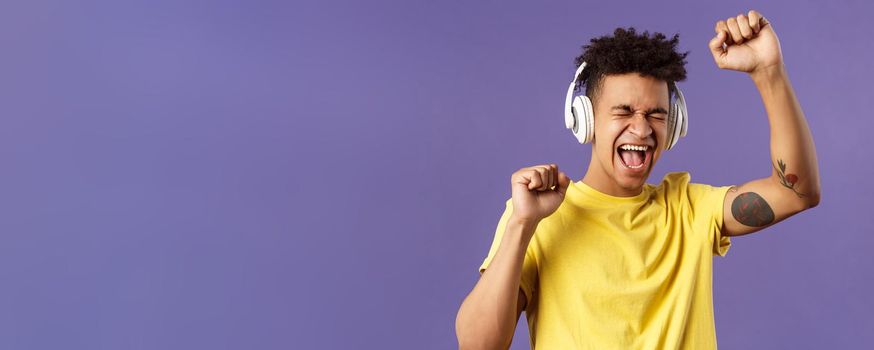 Close-up portrait of cheerful, happy young dancing guy lift hand up singing along, close eyes and smiling upbeat as listening awesome song in headphones, enjoying music, purple background.