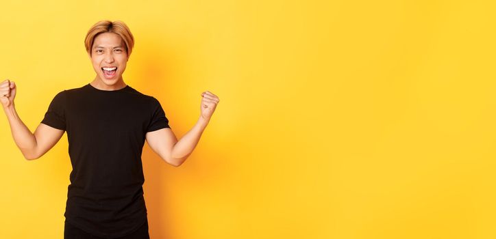 Portrait of happy attractive asian man rejoicing, raising hands up and shouting yes, standing pleased over yellow background.