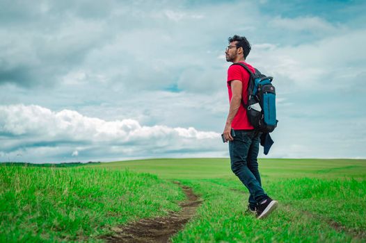 Side view of backpacking man on a beautiful road in the countryside. Young explorer man walking in a green field, Backpacker man walking in a beautiful field.