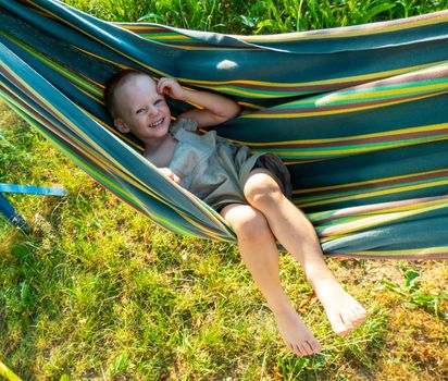 Cute little blond caucasian boy having fun with multicolored hammock in backyard or outdoor playground. Summer active leisure for kids. Child on hammock. Activities and fun for children outdoors. High quality photo