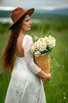 A middle-aged woman in a white dress and brown hat stands on a green field and holds a basket in her hands with a large bouquet of daisies. In the background there are mountains and a lake