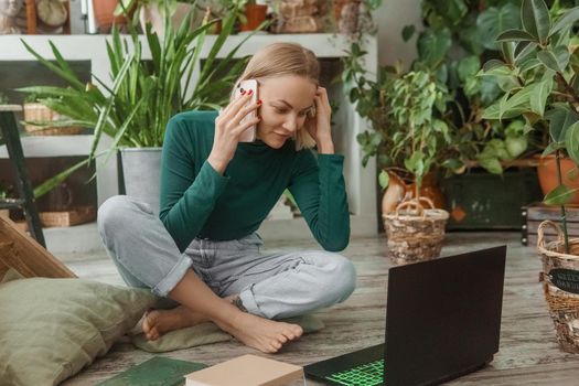 A blonde woman in a room with a lot of green indoor plants is working on a laptop. The concept of biophysical design in the interior. Work from home, work as a freelancer.