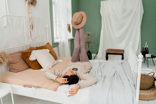 Lifestyle concept. A young middle-aged woman in a sweater lies on the bed on her back, laughs and holds a brown hat on her feet