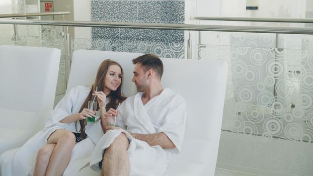Young happy married couple is relaxing sitting in modern spa salon with cocktail glasses and chatting. Romantic relationship, relaxation and wellness concept.