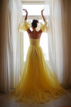 A woman's silhouette in a golden luxurious dress against the background of a window holds a curtain with her hands. Elegant lady in a yellow long silk dress with bare back, back view