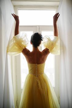 A woman's silhouette in a golden luxurious dress against the background of a window holds a curtain with her hands. Elegant lady in a yellow long silk dress with bare back, back view