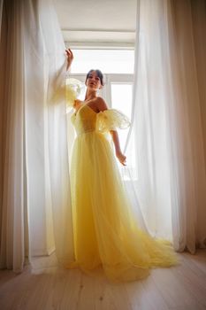 A woman's silhouette in a golden luxurious dress against the background of a window holds a curtain with her hands. Elegant lady in a yellow long silk dress with bare back, back view