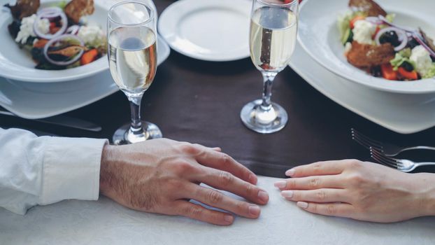 Close-up shot of young lovers holding hands on table at romantic dinner in classy restaurant. Sparking champagne glasses, flatware and food in background.