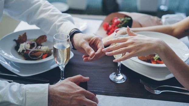 Close-up shot of beautiful ring being placed on manicured female finger during marriage proposal at romantic dinner in restaurant. Engagement and fine dining concept.
