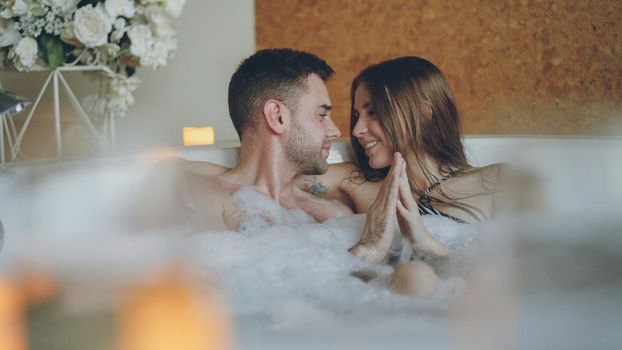 Newly married attractive couple is relaxing in jacuzzi kissing, touching hands, talking and laughing. Romantic relationship, passionate honeymoon and wellness concept.