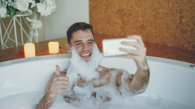 Handsome cheerful guy is taking selfie in bathtub with soap foam on his beard using smart phone. He is laughing and making gestures with his hand posing and having fun.