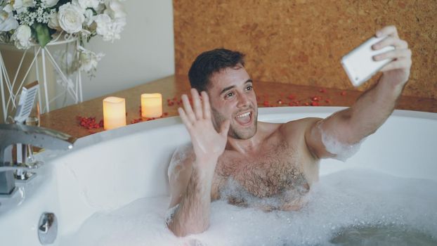 Young handsome man popular blogger is recording video in hot tub in day spa using smartphone for his blog. Burning candles, champagne glass and flowers are visible.