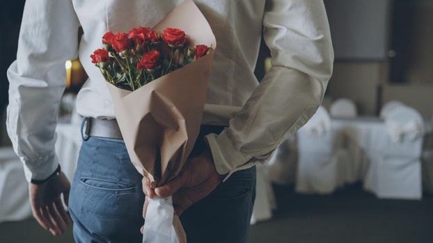 Loving man is hiding red roses in craft paper behind his back bringing beautiful bouquet for his date in restaurant. Flowers, romantic relationship and dating concept.