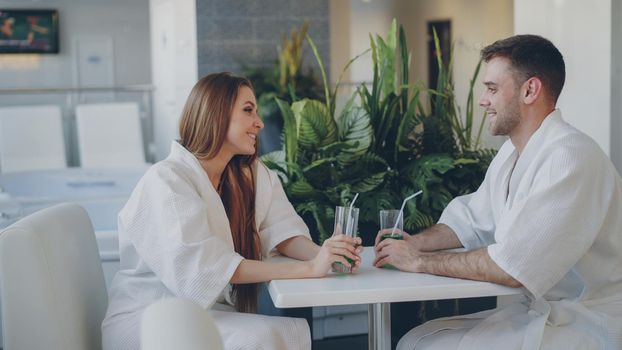 Cheerful young people are talking and drinking while sitting at table in day spa. Comfortable white furniture and green plant are visible.