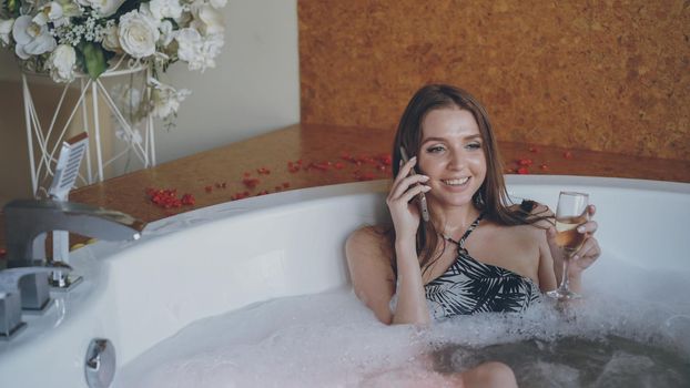 Pretty young woman in bathing suit is talking on mobile phone and drinking champagne while taking bubbling bath in spa salon. Communication, people and relaxation concept.