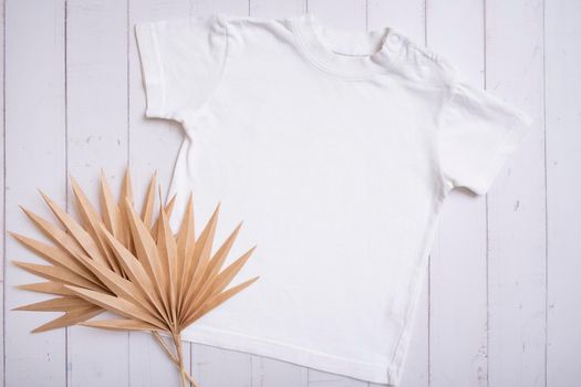 White children's t-shirt mockup for logo, text or design on wooden background with palm leaves top view.