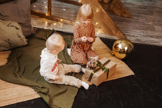 Little brother and sister play on Christmas eve in a beautiful house decorated for the New Year holidays. Children are playing with a Christmas gift. Scandinavian-style interior with live fir trees and a wooden staircase.