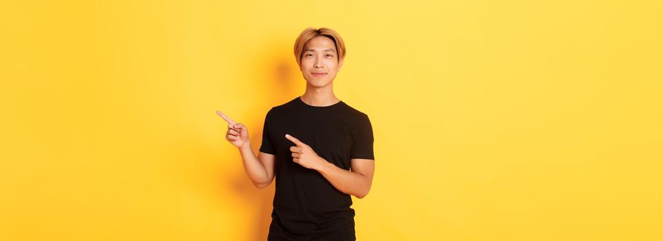 Smiling asian male student with blond hair, pointing fingers right, showing logo, standing yellow background.