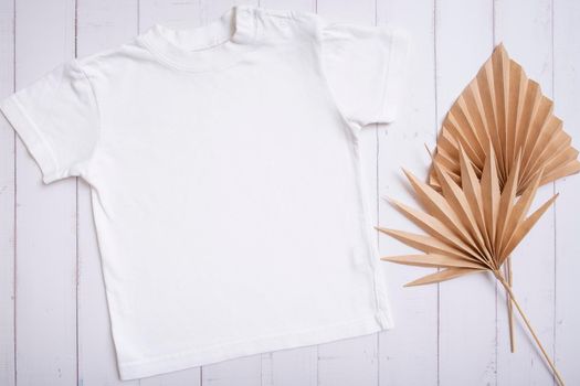 White children's t-shirt mockup for logo, text or design on wooden background with palm leaves top view.