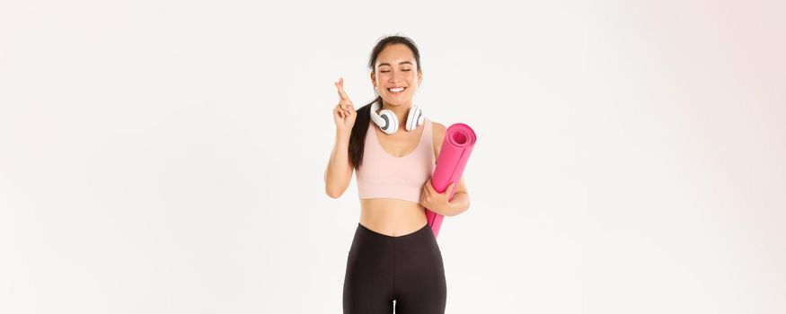 Sport, wellbeing and active lifestyle concept. Hopeful cute asian girl dreaming of going to gym after covid-19 quarantine, holding rubber mat and cross fingers while making wish, pleading.
