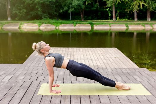A woman in a gray top and leggings, on a wooden platform by a pond in the park in summer, does yoga, on a green mat, performing a reverse plank. Copy space