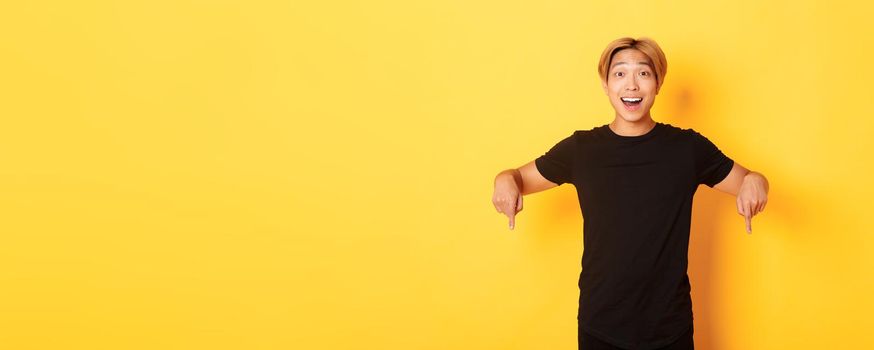 Portrait of excited smiling asian guy with blond hair, pointing fingers down, showing banner and looking happy, yellow background.