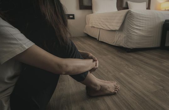 Depressed and stressed woman sitting on vinyl tiles floor near bed in hotel bedroom. Sad woman sitting in the bedroom. Girl with mental health problems. Unhappy life. World Mental Health Day concept.