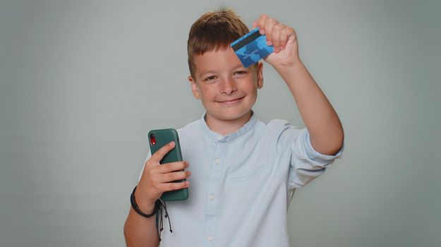 Sincere toddler boy customer using credit bank card and smartphone while transferring money, purchases online shopping, payment. Finance, internet. Young teenager child isolated on gray background
