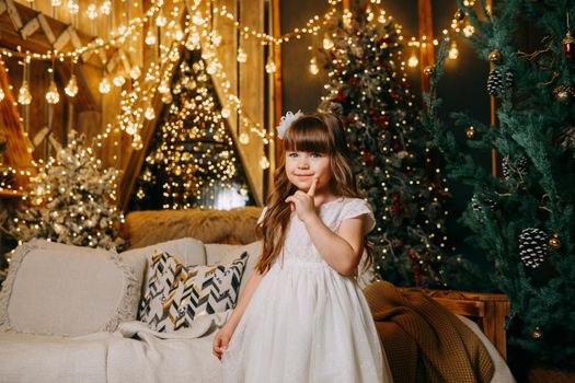 A girl in a festive light dress with a gift in her hands next to a Christmas tree, lights of garlands in the background. The concept of New Year holidays.