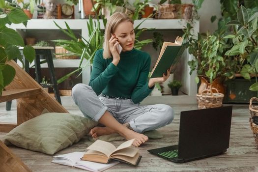 A blonde woman in a room with a lot of green indoor plants is working on a laptop. The concept of biophysical design in the interior. Work from home, work as a freelancer.