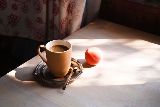 A cup of coffee on vintage matal plate on white wooden table with a peach in the rustic kitchen, summer morning concept, selective focus, copy space.