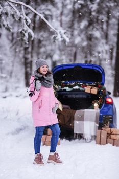 A woman in a winter snow-covered forest in the trunk of a car decorated with Christmas decor. A female photographer holds a camera in her hands.