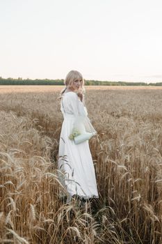 A blonde woman in a long white dress walks in a wheat field. The concept of a wedding and walking in nature.