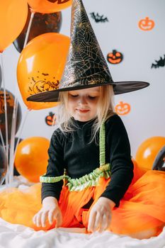 Children's Halloween - a girl in a witch hat and a carnival costume with airy orange and black balloons at home. Ready to celebrate Halloween.