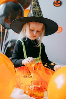 Children's Halloween - a girl in a witch hat and a carnival costume with airy orange and black balloons at home. Ready to celebrate Halloween.