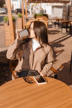 A stylish brunette woman is sitting at a table in an outdoor cafe with coffee and a phone. Autumn walk.