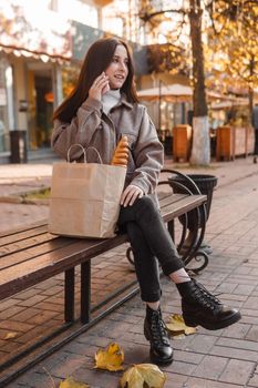 A stylish brunette woman walks around the autumn city. The brunette is sitting on a bench outside with a glass of coffee and a phone, holding a craft bag with a baguette.