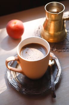 A cup of coffee on vintage metal plate with yellow copper cezve and a peach on white wooden table, summer morning concept, selective focus.